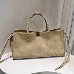May include: A beige suede tote bag with two top handles and a rectangular shape. The bag has a silver metal clasp and long, thin straps on each side. The bag is sitting on a white surface.