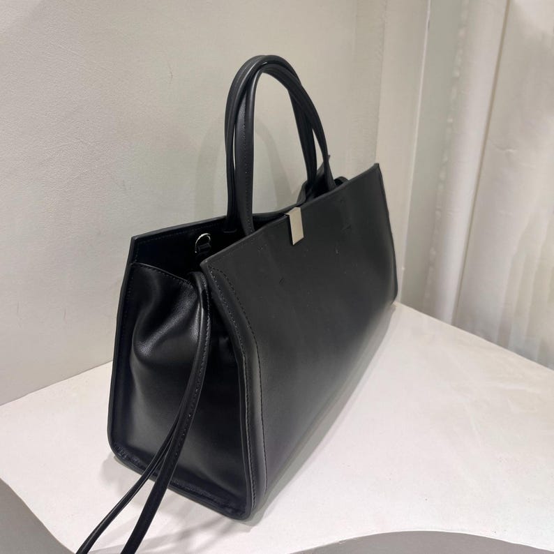May include: A black handbag with a rectangular shape and two handles. The bag has a silver-colored clasp and a minimalist design. The bag is made of a smooth material and has a structured silhouette.
