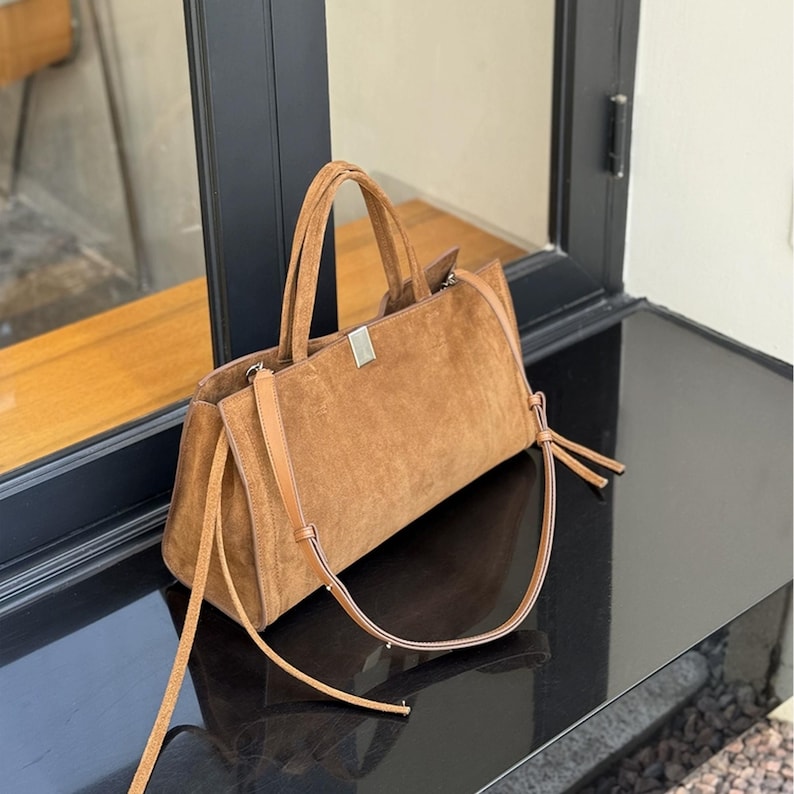 May include: A brown suede handbag with a rectangular shape. The bag has two handles and a long, adjustable shoulder strap. A silver-colored metal bar is on the top of the bag. The bag is sitting on a black surface.