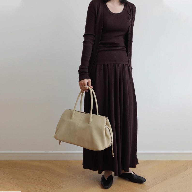 K&ouml;nnte beinhalten: Eine Frau tr&auml;gt ein dunkelburgunderfarbenes Outfit, bestehend aus einer lang&auml;rmligen Strickjacke, einem Oberteil mit Rundhalsausschnitt und einem flie&szlig;enden Rock. Sie h&auml;lt eine gro&szlig;e, cremefarbene Lederhandtasche mit zwei Griffen. Das Outfit wird durch schwarze Schuhe erg&auml;nzt.