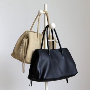K&ouml;nnte beinhalten: Zwei Tragetaschen h&auml;ngen an einem wei&szlig;en Kleiderst&auml;nder. Eine Tasche ist aus hellbeige Wildleder mit Rei&szlig;verschluss und Fransen. Die andere ist eine schwarze Ledertasche mit langen Henkeln und strukturierter Oberfl&auml;che.