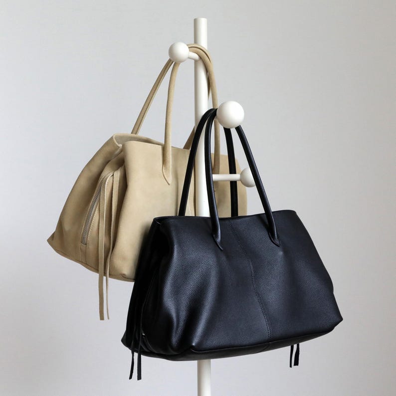 K&ouml;nnte beinhalten: Zwei Tragetaschen h&auml;ngen an einem wei&szlig;en Kleiderst&auml;nder. Eine Tasche ist aus hellbeige Wildleder mit Rei&szlig;verschluss und Fransen. Die andere ist eine schwarze Ledertasche mit langen Henkeln und strukturierter Oberfl&auml;che.