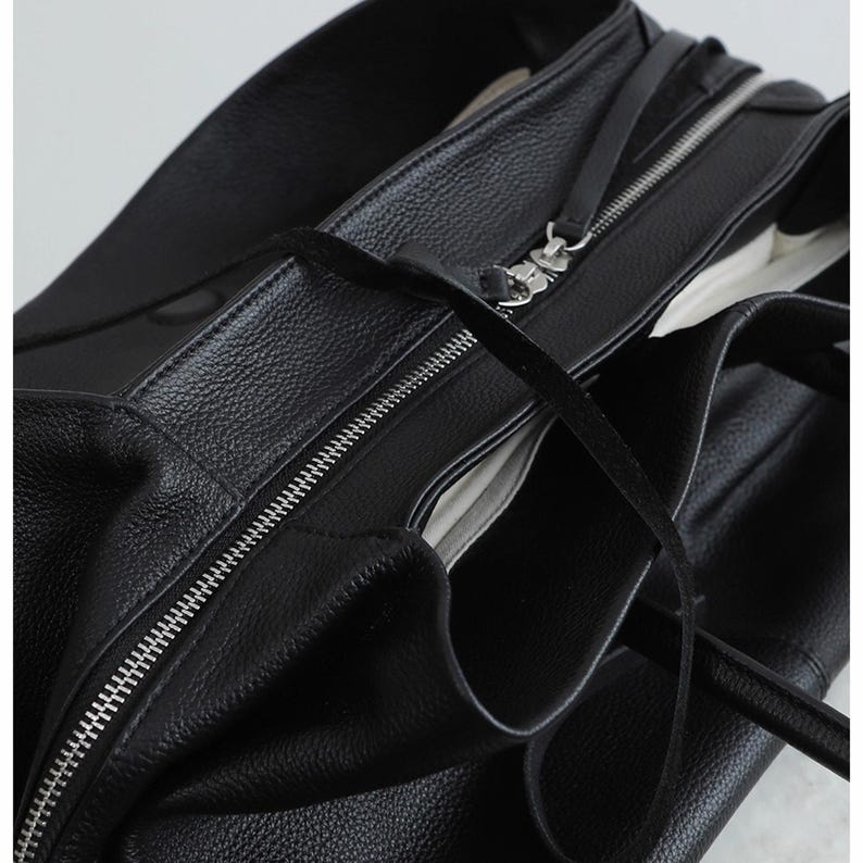 K&ouml;nnte beinhalten: Schwarze Ledertasche mit silbernem Rei&szlig;verschluss. Die Tasche hat eine strukturierte Oberfl&auml;che und einen langen, d&uuml;nnen Riemen. Das Innere der Tasche ist sichtbar und zeigt ein helleres Futter. Die Tasche ist offen.