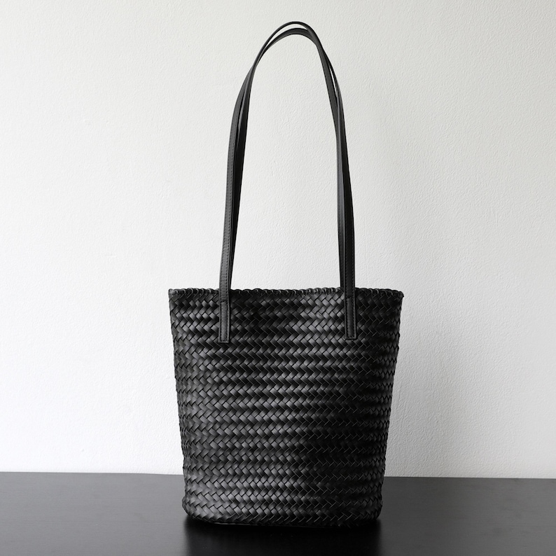 Peut inclure: Sac fourre-tout noir tiss&eacute; avec de longues anses fines. Le sac a une forme cylindrique et est fait d'un mat&eacute;riau tiss&eacute; serr&eacute;. Les anses sont fix&eacute;es en haut du sac et sont faites d'un mat&eacute;riau lisse et uni.