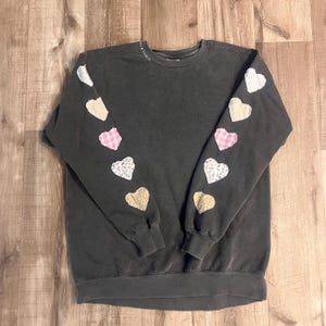 May include: A dark gray sweatshirt with a crew neck and long sleeves. The sleeves have a row of five patchwork hearts in different colors and patterns, including pink gingham, floral print, and beige.