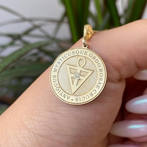 May include: A gold pendant necklace with a round charm featuring a triangle with an ankh symbol inside. The charm is inscribed with the text "MYSTICUSQUE ORDO ROSAE CRUCIS ANTIOQUUS".