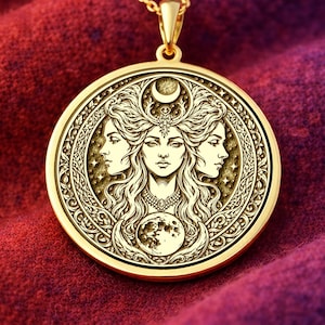 Può includere: Collana con pendente color oro con un design dettagliato di una donna con tre volti, una luna crescente e una luna piena. Il pendente è appeso a una delicata catena d'oro. La collana è esposta su un tessuto strutturato rosso e viola.