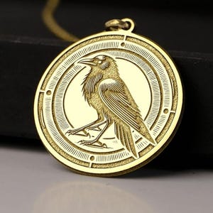May include: A gold-colored pendant necklace featuring a detailed engraving of a raven. The raven is centered within a circular frame with an intricate border design. The pendant hangs from a delicate gold chain, set against a dark background.
