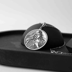 May include: A silver pendant necklace featuring a circular design with a woman's face on one side and a skeleton on the other. The pendant is on a black surface.