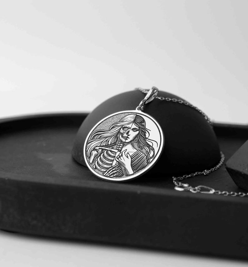 May include: A silver pendant necklace featuring a circular design with a woman's face on one side and a skeleton on the other. The pendant is on a black surface.