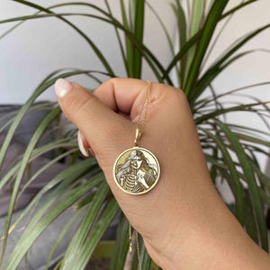 May include: Gold necklace with a round pendant featuring a detailed image of a woman with a skeleton on one side of her face. The pendant is on a gold chain.
