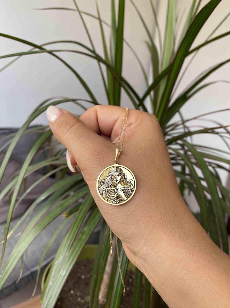 May include: Gold necklace with a round pendant featuring a detailed image of a woman with a skeleton on one side of her face. The pendant is on a gold chain.