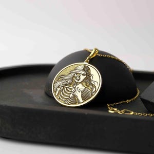May include: Gold pendant necklace featuring a round charm with an engraved image of a woman's face and a skeleton. The woman's face is on the right side of the charm and the skeleton is on the left side.