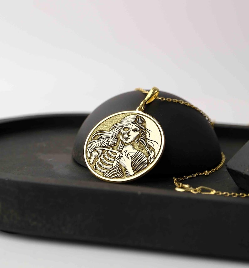 May include: Gold pendant necklace featuring a round charm with an engraved image of a woman's face and a skeleton. The woman's face is on the right side of the charm and the skeleton is on the left side.