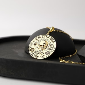 May include: Gold pendant necklace with a round charm featuring a skull, crossbones, hourglass, compass, and flowers. The charm has the text "Memento Mori" and "Amor Fati" around the edge.