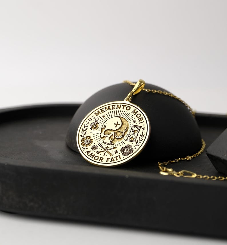 May include: Gold pendant necklace with a round charm featuring a skull, crossbones, hourglass, compass, and flowers. The charm has the text "Memento Mori" and "Amor Fati" around the edge.