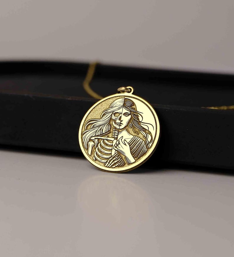 May include: A gold-toned pendant necklace features a round charm with a detailed image of a woman and a skeleton, their bodies intertwined. The woman's face is visible on the right side of the charm, while the skeleton's ribs are visible on the left side.
