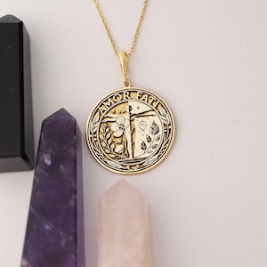 May include: Gold necklace with a round pendant featuring the words "Amor Fati" and a stylised image of a person with outstretched arms, a flower, and leaves.