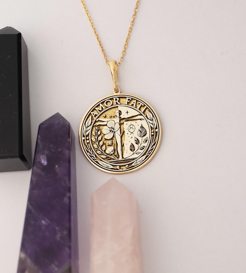 May include: Gold necklace with a round pendant featuring the words "Amor Fati" and a stylised image of a person with outstretched arms, a flower, and leaves.