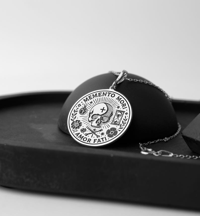 May include: Silver pendant necklace with a circular design featuring a skull, crossbones, hourglass, compass, and flowers. The pendant reads "Memento Mori" and "Amor Fati".