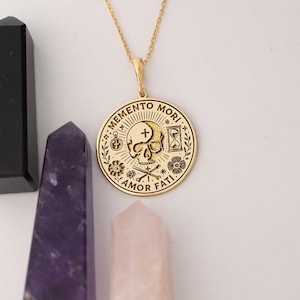 May include: Gold necklace with a round pendant featuring a skull with a cross, an hourglass, and the words "Memento Mori" and "Amor Fati".