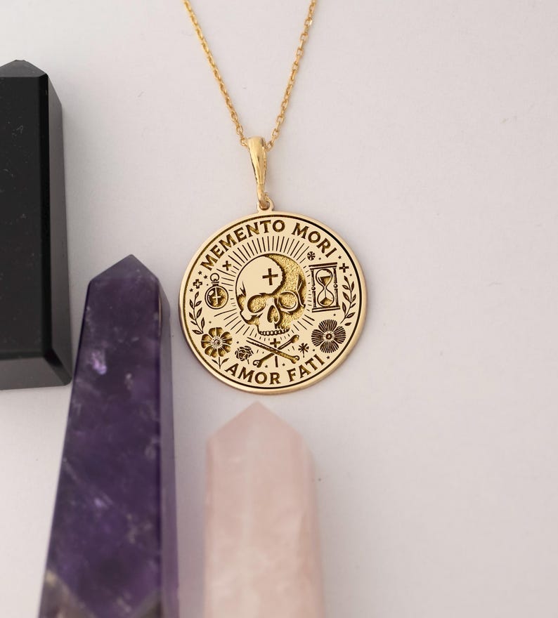 May include: Gold necklace with a round pendant featuring a skull with a cross, an hourglass, and the words "Memento Mori" and "Amor Fati".