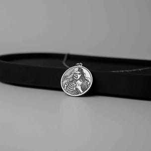 May include: Silver pendant necklace featuring a circular design with a woman's face on one side and a skeleton on the other. The woman's face is partially obscured by the skeleton.