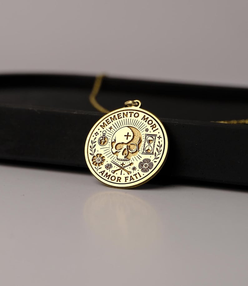 May include: Gold necklace with a round pendant featuring a skull with a cross, an hourglass, and the words "Memento Mori" and "Amor Fati".