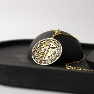 May include: A gold pendant necklace with a round charm featuring a stylised figure with outstretched arms surrounded by floral motifs. The charm is inscribed with the words "AMOR FATI".