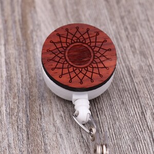 Puede incluir: Un portagafete retráctil blanco con un disco redondo de madera con un diseño de mandala rojo.