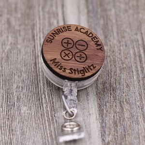 Peut inclure: Un porte-badge en bois avec un cordon rétractable. Le badge porte le texte "Sunrise Academy" et "Miss Stiglitz" avec des symboles mathématiques.