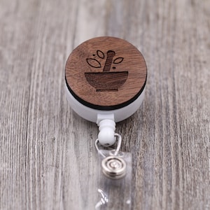 May include: A white and wood-tone retractable badge reel. The wooden top features a carved mortar and pestle design with leaves. The reel has a clear plastic strap and a metal clip for attaching to clothing.