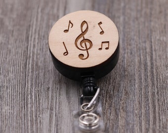 May include: A wooden retractable badge holder with a black and white treble clef and musical notes design. The holder has a black plastic base and a clear plastic retractable cord.