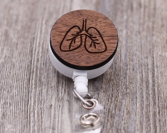 Op de afbeelding: Een ronde, houten badge reel met een lasergeëtst longontwerp. De donkerbruine houten bovenkant contrasteert met de witte behuizing van de reel. Het heeft een transparante plastic band en een metalen clip. Praktisch accessoire.