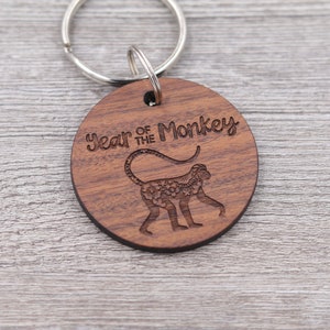 Puede incluir: Un llavero de madera con fondo marrón y un llavero plateado. El llavero presenta una silueta de mono con el texto "Year of the Monkey" encima.