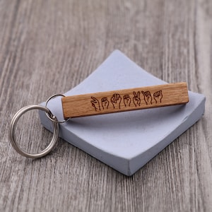 May include: Wooden keychain with a silver key ring. The keychain has a woodgrain pattern and is engraved with a series of hand signs in American Sign Language.