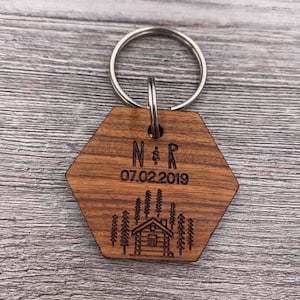 May include: Wooden hexagon keyring with a cabin and trees design. The keyring is engraved with the initials "N & R" and the date "07.02.2019".