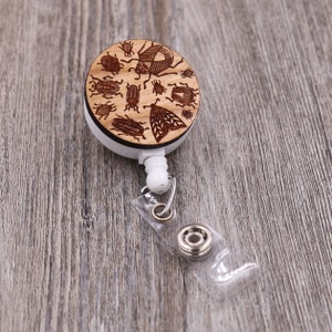May include: A wooden retractable badge holder with a design of various insects, including beetles and butterflies. The holder is white with a clear plastic strap.