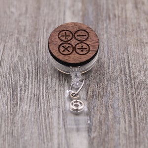 Peut inclure: Un porte-badge en bois avec un cercle en bois brun sur lequel sont gravés les symboles mathématiques d'addition, de soustraction, de multiplication et de division. La bobine est en plastique transparent et possède un cordon rétractable argenté.