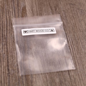K&ouml;nnte beinhalten: Eine durchsichtige Plastikt&uuml;te mit einem wei&szlig;en Etikett, auf dem "HART WOOD CO." mit einem Herz und einem Kiefernbaum-Symbol steht.