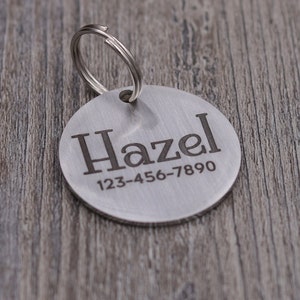 Könnte beinhalten: Ein runder, gebürsteter Metall-Haustieranhänger mit dem Namen "Hazel" und einer Telefonnummer, die in dunkler Schrift eingraviert ist. Der Anhänger ist an einem silbernen Spaltring befestigt. Der Hintergrund ist eine Holzoberfläche.