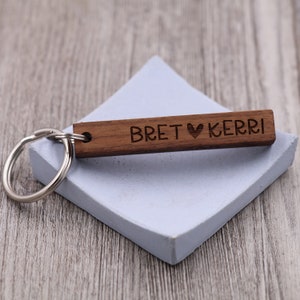 May include: A wooden keychain with the names "Bret" and "Kerri" engraved on it, separated by a heart symbol. The keychain is brown and has a silver key ring.