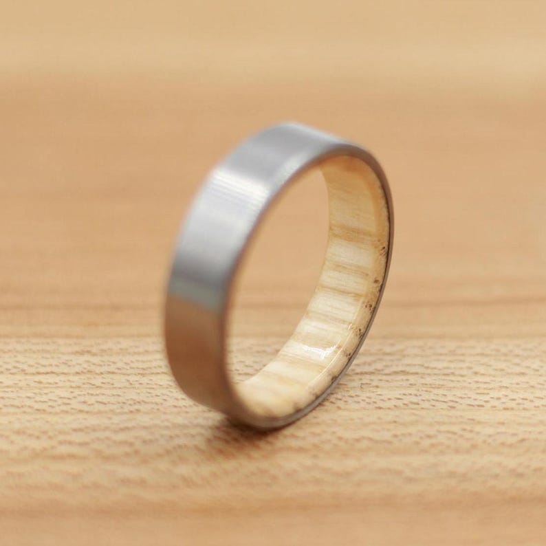 May include: A silver-colored ring with a light-colored wooden interior. The ring has a flat, brushed metal exterior and a natural wood grain interior. The ring is likely a wedding band or a fashion accessory.