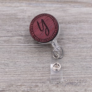 May include: A wooden retractable badge holder with a dark red finish and a white circle around the edge. The circle has the letter "Y" engraved in the centre. The holder is attached to a clear plastic retractable cord and a silver clip.
