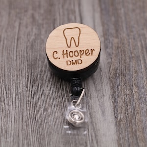 Puede incluir: Portagafete retráctil de madera con un retractor negro y un clip de plástico transparente. La parte de madera es redonda y presenta un icono de diente y el texto "C. Hooper DMD".