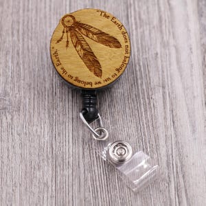 May include: A wooden badge reel with a dreamcatcher and feather design. The text around the edge reads, "The Earth does not belong to us; we belong to the Earth." The reel has a black cord and a clear plastic clip.