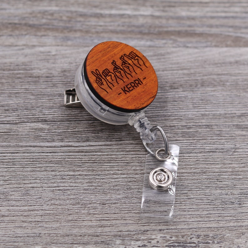 May include: A wooden badge reel with a clear plastic casing. The wooden disc features a design of hands signing the name "KERRI" in American Sign Language.