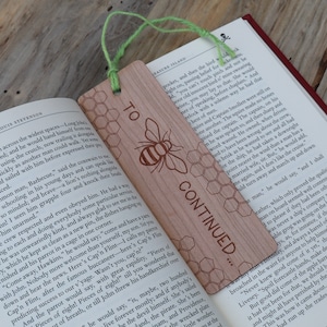 May include: A wooden bookmark with a honeybee design and the text "To be Continued..."  The bookmark is light brown with a green string attached to the top.