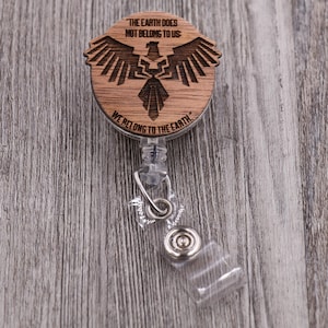 May include: A wooden badge reel with a laser-cut design of an eagle and the text "THE EARTH DOES NOT BELONG TO US" and "WE BELONG TO THE EARTH". The reel has a clear plastic strap and a metal clip.