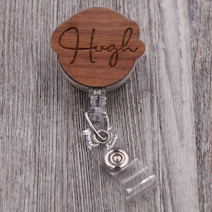 May include: A wooden name badge reel with the name "Hugh" engraved in a cursive font. The badge reel has a clear plastic strap and a metal clip. The wooden portion is a light brown color.